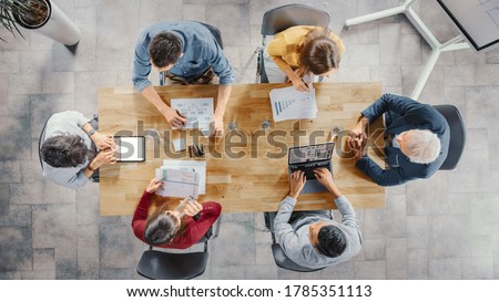Startup Meeting Room: Team of Entrepreneurs sitting at the Conference Table Have Discussions, Solve Problems, Use Digital Tablet, Laptop, Share Documents with Statistics, Charts. Top View