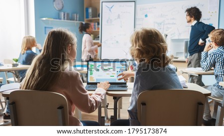 Elementary School Science Class: Over the Shoulder Little Boy and Girl Use Laptop with Screen Showing Programming Software. Physics Teacher Explains Lesson to a Diverse Class full of Smart Kids