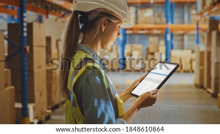 Professional Female Worker Wearing Hard Hat Uses Digital Tablet Computer with Inventory Checking Software in the Retail Warehouse full of Shelves with Goods. Delivery, Distribution Center.