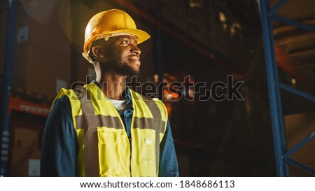Handsome Professional Worker Wearing Safety Vest and Hard Hat Charmingly Smiling and Looks Into the Distance. In the Background Big Warehouse with Shelves full of Delivery Goods. Medium Portrait