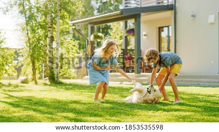 Two Kids Have fun with Their Handsome Golden Retriever Dog on the Backyard Lawn. They Pet, Play, Scratch it. Happy Pedigree Dog Holds Toy ball in Jaws. Idyllic Suburb House in the Summer