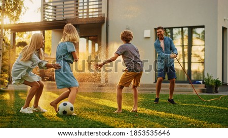Happy Family of Four Playing with Garden Water Hose, Spraying Each Other. Mother, Father, Daughter and Son Have Fun Playing Games in the Backyard Lawn of Idyllic Suburban House on Sunny Summer Day