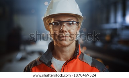 Portrait of a Professional Heavy Industry Engineer Worker Wearing Uniform, Glasses and Hard Hat in a Steel Factory. Beautiful Female Industrial Specialist Standing in Metal Construction Facility.