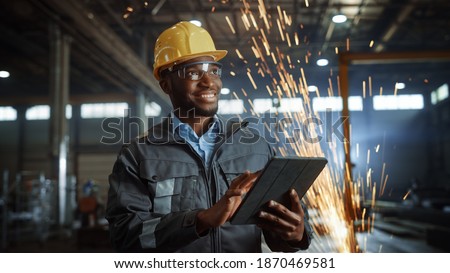 Professional Heavy Industry Engineer Worker Wearing Safety Uniform and Hard Hat Uses Tablet Computer. Smiling African American Industrial Specialist Standing in a Metal Construction Manufacture. Foto d'archivio © 