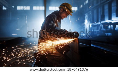 Heavy Industry Engineering Factory Interior with Industrial Worker Using Angle Grinder and Cutting a Metal Tube. Contractor in Safety Uniform and Hard Hat Manufacturing Metal Structures. 商業照片 © 