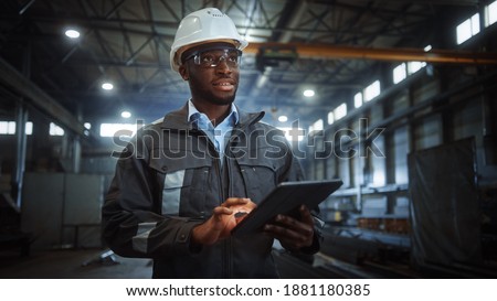 Professional Heavy Industry Engineer Worker Wearing Safety Uniform and Hard Hat Uses Tablet Computer. Smiling African American Industrial Specialist Walking in a Metal Construction Manufacture.
