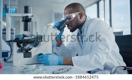 Medical Development Laboratory: Black Male Scientist Looking Under Microscope, Inspecting Petri Dish. Professionals Working in Advanced Scientific Lab doing Medicine, Vaccine, Biotechnology Research