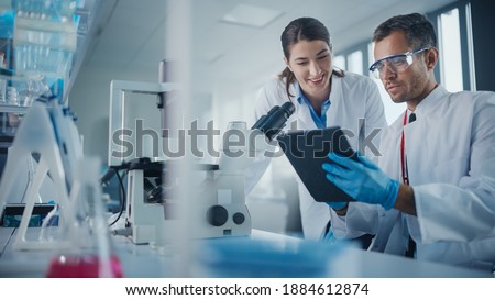 Modern Medical Research Laboratory: Portrait of Two Scientists Working, Using Digital Tablet, Analyzing Samples, Talking. Advanced Scientific Pharmaceutical Lab for Medicine, Biotechnology Development