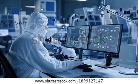 Electronics Factory Cleanroom: Engineer Scientist in Coveralls Works on Computer, Screen Shows Infographics and Software System Control UI, Developing Electronics for Medical Electronics