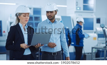 Chief Engineer and Project Manager Wearing Safety Vests and Hard Hats, Use Laptop in Modern Factory, Talking, Optimizing Production Line. Industrial Facility: Professionals Working on Machinery Stockfoto © 