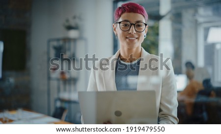 Modern Office: Portrait of Beautiful Creative Specialist with Short Pink Hair Standing, Holding Laptop Computer. Working on App Design, Data Analysis, Plan Strategy for Social Media Disruption