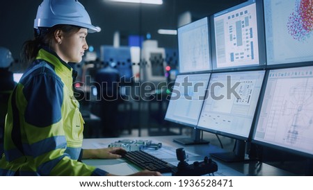 Industry 4.0 Modern Factory: Female Facility Operator Controls Workshop Production Line, Uses Computer with Screens Showing Complex UI of Machine Operation Processes, Controllers, Machinery Blueprints
