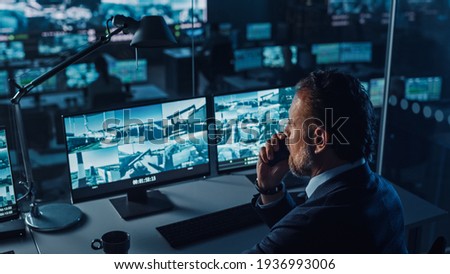 Male Officer Works on a Computer with Surveillance CCTV Video in a Harbour Monitoring Center with Multiple Cameras on a Big Digital Screen. Employee Uses Radio to Give an Order or Report.