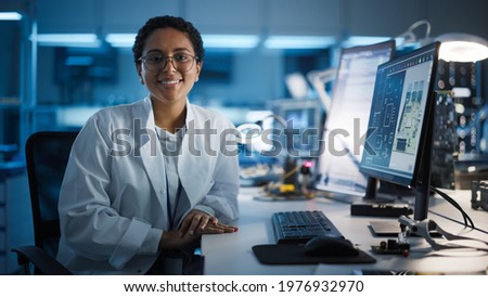 Beautiful Black Latin Woman Wearing Glasses Smiling Charmingly Looking at Camera. Young Intelligent Female Scientist Working in Laboratory. Technological Laboratory in Bokeh Blue as Background