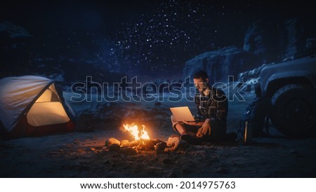 Night Tent Camping in Canyon: Male Traveler Uses Laptop Computer Sitting by Campfire. Man on Digital Remote Work, e-shopping, ecommerce, Using Internet, Social Media Posting on Vacation Trip