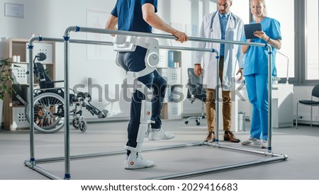 Modern Hospital Physical Therapy: Patient with Injury Walks Wearing Advanced Robotic Exoskeleton. Physiotherapy Rehabilitation Scientists, Engineers use Tablet Computer to Help Disabled Person