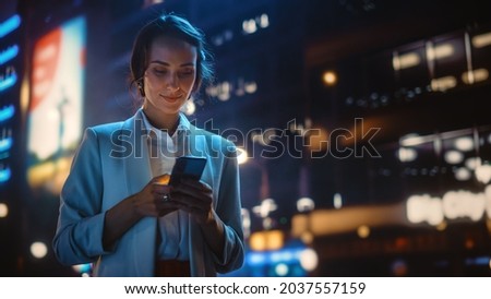 Photo of Beautiful Young Woman Using Smartphone Walking Through Night City Street Full of Neon Light. Portrait of Gorgeous Smiling Female Using Mobile Phone.