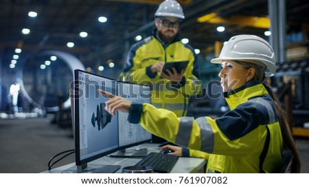 Inside the Heavy Industry, Factory Female Industrial Engineer Works on Personal Computer She Designs 3D Engine Model, Her Male Colleague Talks with Her and Uses Tablet Computer. Low Angle Shot. 商業照片 © 