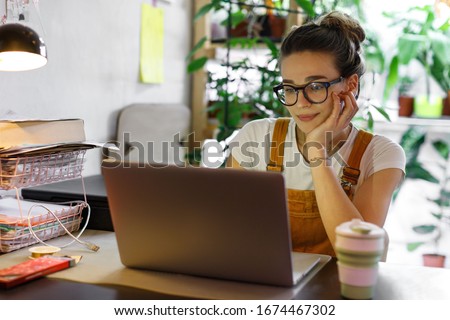 Young female gardener in glasses using laptop, communicates on internet with customer in home garden/greenhouse, reusable coffee/tea mug on table.Cozy office workplace, remote work, E learning concept 商業照片 © 