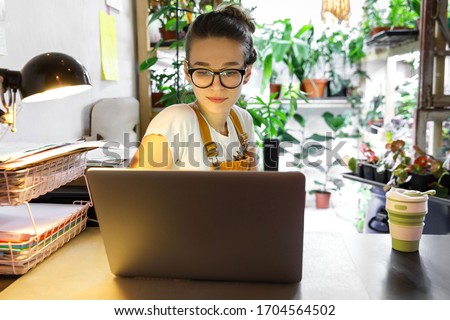 European female gardener in glasses using laptop, scrolling through social networks, reads news, coffee/tea mug on table, home garden/greenhouse on background. Cozy workplace, remote work