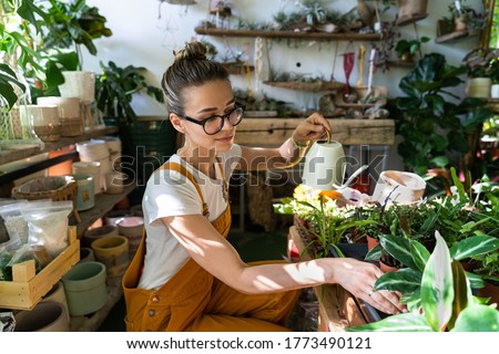 Woman gardener in orange overalls watering potted houseplant in greenhouse surrounded by plants and pots, using white watering can metal. Home gardening, love of plants and care. Small business. 