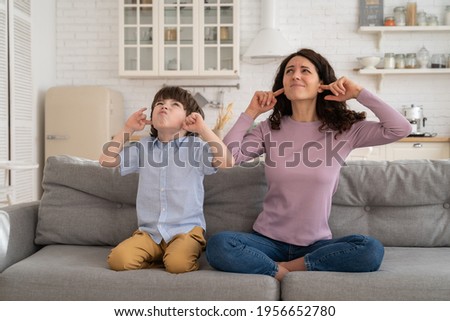 Frowning mom and son sit on couch with closed eyes and cover ears from noisy music or fight sounds from neighbors. Young female parent and kid tired of drilling noise not listen plug ears with fingers