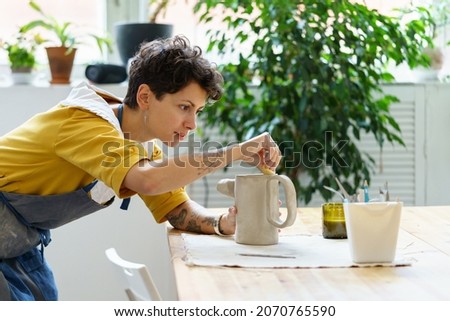 Young female artisan concentrated on modeling jug from raw clay while pottery lesson or workshop in creative studio. Woman ceramic business owner making craft for sale in handmade potter retail store Stockfoto © 