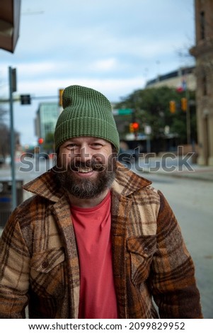 A happy bearded man wearing a green knitted beanie and a brown plaid coat smiles for a portrait in a downtown setting on a sunny day. Stock foto © 