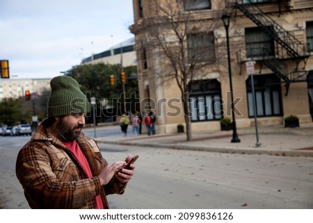 A bearded man wearing a green knitted beanie uses a smartphone in the city in this technology lifestyle concept.  The cell phone uses data and a wireless network as he communicates digitally. Stock foto © 