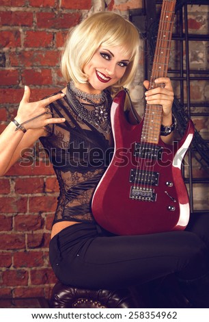 Portrait of a cool glam rock blonde sexy woman in black lingerie with red guitar on a brick wall background