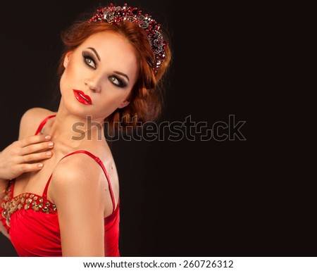 Amazing beautiful red hair woman with party make up in red dress with golden crystals and luxury princess tiara dreaming posing tender on black background with copyspace