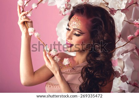 Fashion Art Beauty Portrait. Beautiful woman with fantasy creative make up in spring garden holding flowering sakura branch. Pink background. Spring time concept