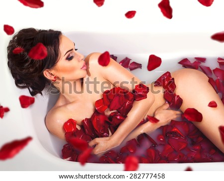 Sexy beautiful naked brunette woman laying in a bath with milk and red romantic falling  rose petals. Luxury spa and skin care