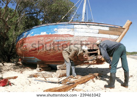 Two men repair a hole in the hull of a damaged wooden fishing boat in rural Africa.