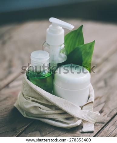 Cosmetic bottle container with green herbal leaves in small organic cotton bag on wooden background. Toned image