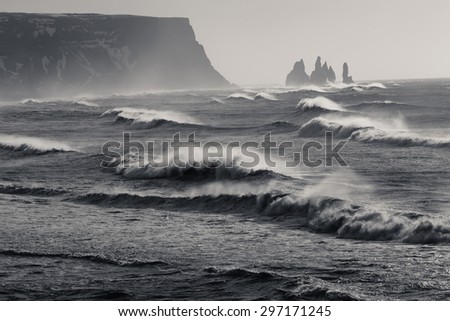 Black and White of Monster waves crashing with jagged rocks and cliffs in the distance on DyrhÃ³laey beach in Iceland.