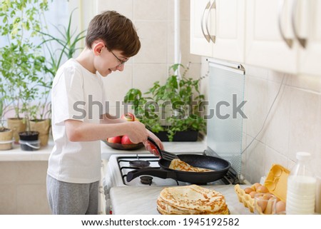 teen boy bakes (fries) pancakes in the kitchen. Cooking at home. Teenager boy is learning to cook. Kid boy 12 years old makes crepes by himself on frying pan. Independent child. Novice young chef