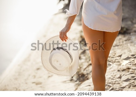 Young woman on a beach holdind white hat. Summer holiday concept travel. Young woman weared in White shirt on blured nature background. Summer story. Legs close up Imagine de stoc © 