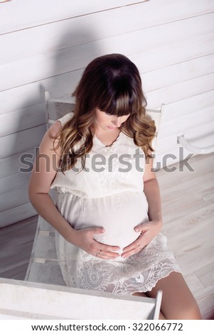 Pregnant woman looks at the stomach. Romantic hairstyle, white lacy dress , white background, wooden furniture