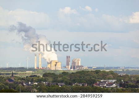 View from a slag heap over rural landscape to a distant steaming coal-fired power station surrounded by wind turbines.