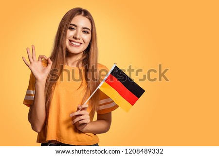 A beautiful young woman holds in her hands the flag of Germany and shows the OK sign. Exchange student, learn Germanic language. Tourist traveling. Yellow, orange background. Football fan. ストックフォト © 