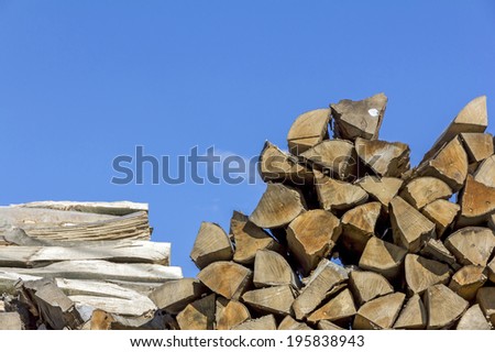 Logs of and boards of wood of different shapes, sizes and kinds piled together in saw-mill with blue sky above in background