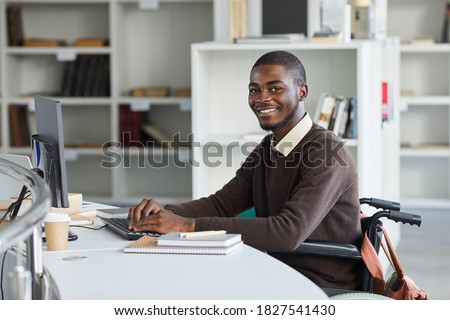 Portrait of disabled African-American man using computer and smiling at camera while studying in college library, copy space