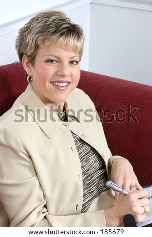 Woman in business suit writing in her datebook, sitting on couch with folders and briefcase nearby.