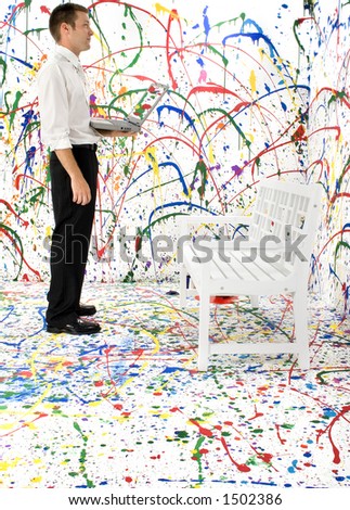 Attractive young business man standing with laptop in front of white bench on paint splattered background.  Looking at wall. Full body.