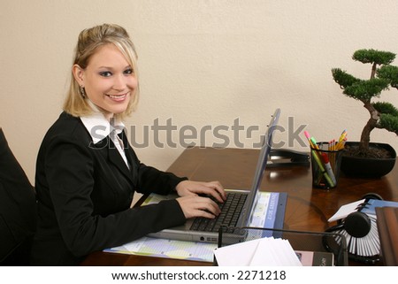 Woman at office desk working on laptop.  Smiling at camera.
