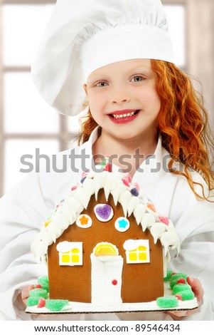 Adorable girl child in chef uniform holding a ginger bread, gingerbread house over white background.