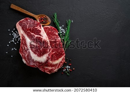 Two Raw beef rib eye steak prepared for grilling with seasonings and herbs on a dark textured background. Fresh meat steak, top view