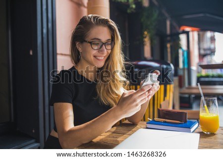 Happy smiling gorgeous woman in fashionable glasses reading pleasant text message on mobile phone while sitting in coffee shop during recreation time. Joyful female watching video on cellphone 