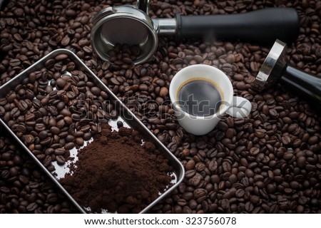 A cup of hot and fresh espresso coffee lay on many roast coffee beans also see an espresso machine group head, coffee tamper and ground coffee lay on many roasted coffee beans.